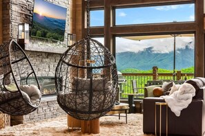 A room with a stone fireplace, a hanging chair, a sofa, and a large window with a mountain view.
