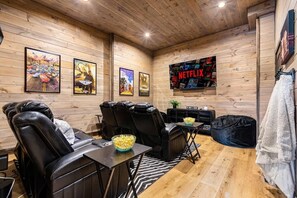 Une pièce aux murs en bois, une affiche de Netflix, des fauteuils inclinables, une table avec un bol de pop-corn et un pouf noir.
