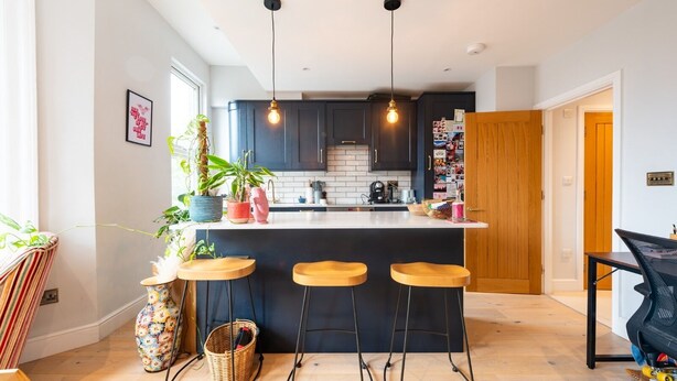 A modern kitchen with dark cabinets, a white countertop, and a central island with stools. There are hanging pendant lights, a potted plant, and a basket on the counter. - VESTO Gauden Road (London)