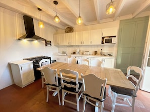 A kitchen with white cabinets, a wooden table, and six white chairs. There is a stove, a microwave, and hanging pendant lights. - La Villa Rouge (Valdivienne)