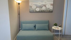 A bedroom with a teal sofa, a painting of dandelions, a bedside table with a lamp, and a small potted plant.