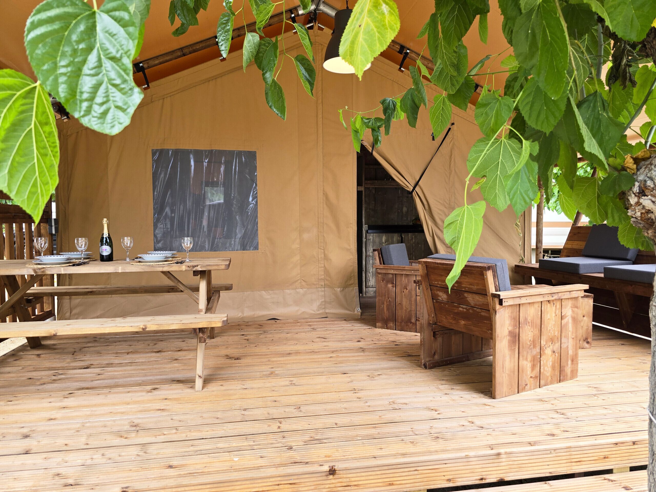 Una terraza de madera con una mesa lista para comer, una zona de estar con bancos y una planta grande y frondosa en lo alto.