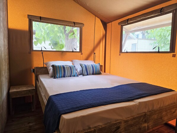 Un dormitorio con una cama de madera, dos ventanas con cortinas, una mesita de noche pequeña y una manta azul.