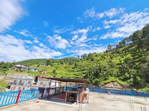 A terrace with a blue railing, a covered seating area, and a view of a hillside with buildings. - Hotel Mountain Paradise Barkot (Barkot)