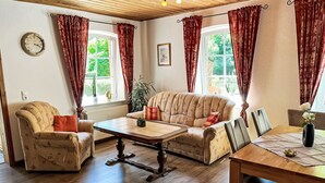 A cozy living room with a sofa, armchair, and a dining table. There are windows with curtains, a clock on the wall, and a plant.