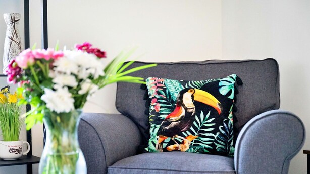 A grey armchair with a tropical bird pillow, a clear vase with flowers, and a mug on a side table.