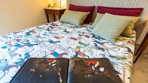 Een bed met een kleurrijke quilt met afbeeldingen van ruiters en paarden, twee kussens en een nachtkastje met een lamp.