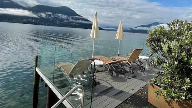 A lakeside deck with lounge chairs and umbrellas, overlooking a calm lake and mountains. - Vacation apartment Brissago (Brissago)