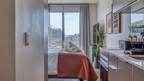 Una habitación de hotel moderna con una cama grande, vistas a la ciudad, una kitchenette y una planta en maceta.