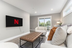 A modern living room with a flat-screen TV mounted on the wall, a tufted leather bench, a white sofa with brown and gray cushions, and a balcony with a view of the outdoors. - The Birches Condotel Unit 2 (Lake Leelanau)