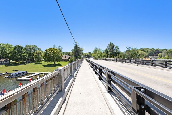 A pedestrian bridge over a river with a clear sky, a dock with boats, and greenery on both sides. - The Birches Condotel Unit 2 (Lake Leelanau)