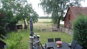 A patio area with a table and chairs, a barbecue grill, and a fenced yard with trees and a brick building in the background.