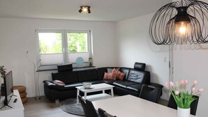A modern living room with a black leather sofa, a white coffee table, and a dining table with chairs. There is a window with blinds, a black pendant light, and a vase with flowers on the dining table.