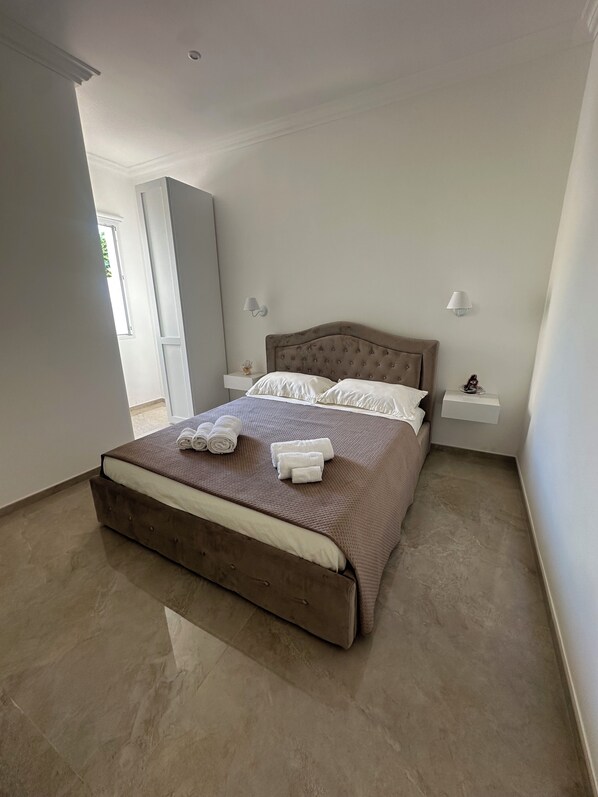 Una cama bien hecha con cabecera marrón y ropa de cama blanca, dos mesitas de noche con lámparas y un gran ventanal que proporciona luz natural.