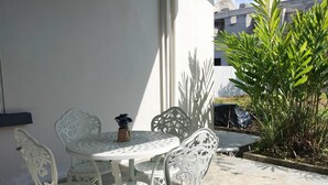 Une terrasse extérieure avec des chaises et une table en métal blanc, entourée d’un mur blanc et d’une grande plante.