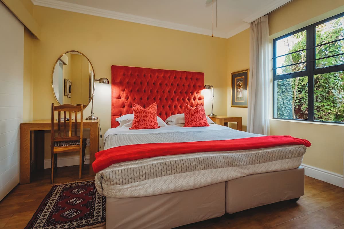 A bedroom with a tufted headboard, a wooden desk, a mirror, a bedside table with lamps, and a large window with a view of greenery.
