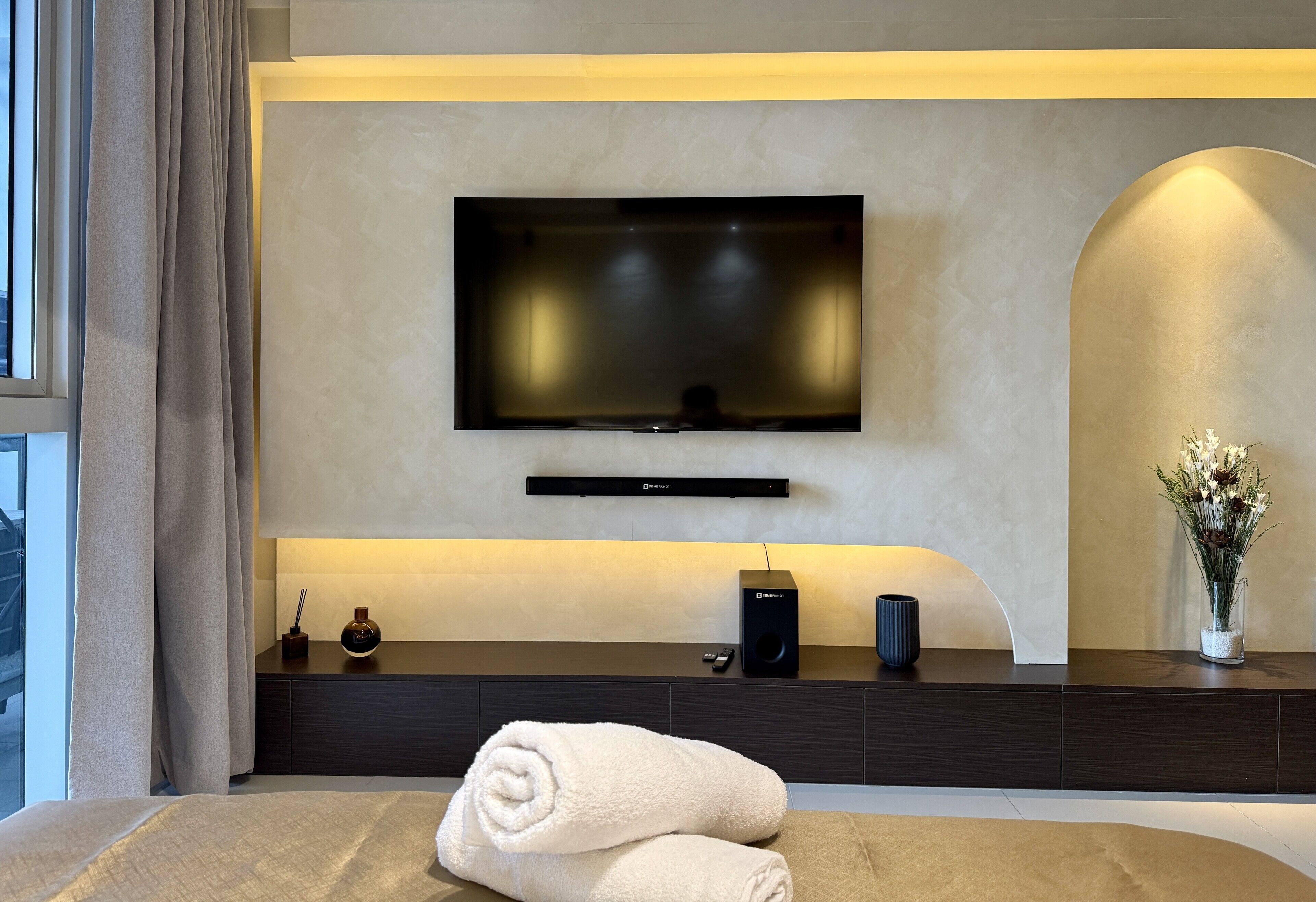 A modern hotel room with a flat-screen TV mounted on the wall, a dark wood console table with a speaker and a vase of flowers, and a neatly folded towel on the bed.