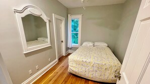 Ein Schlafzimmer mit Holzboden, einem Bett mit gemusterten Textilien, einem Fenster mit Vorhängen und einem Spiegel an der Wand.