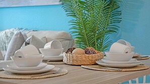 Egy étkezőasztal, melyre fehér tányérok és szőtt alátétek kerültek, egy szőtt kosár dísztárgyakkal, és a háttérben egy nagy, zöld növény.