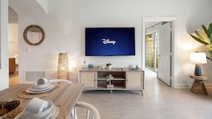 Egy modern nappali, melyben egy síkképernyős televízió látható, amin a Disney logója jelenik meg, egy fa étkezőasztal van terítve tányérokkal és csészékkel, egy fonott lámpa és egy konzolasztal díszíti a teret, melyen dekoratív tárgyak találhatók.