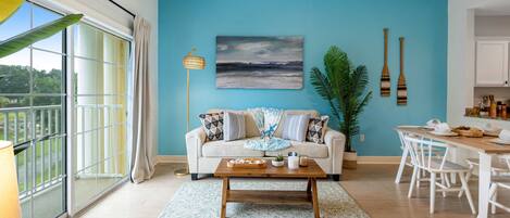 Un salon moderne avec un mur d’accent bleu, une grande fenêtre, un canapé blanc, une table basse en bois et un coin repas avec des chaises blanches.