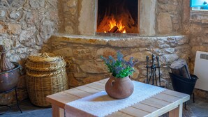 A stone fireplace with a fire, a woven basket, a wooden table with a vase of flowers, and a metal heater. - AKAMANTIDA PLATEA 1. A 3-bedroom country house built in 1900, renovated in 2016. (Pano Arodes)