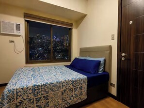 A bedroom with a city view, a bed with a patterned bedspread, a window with blinds, and a door.