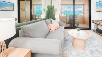 Uma sala de estar moderna com um sofá cinzento, uma mesa de centro redonda de madeira e vista para a praia através de grandes portas de vidro.
