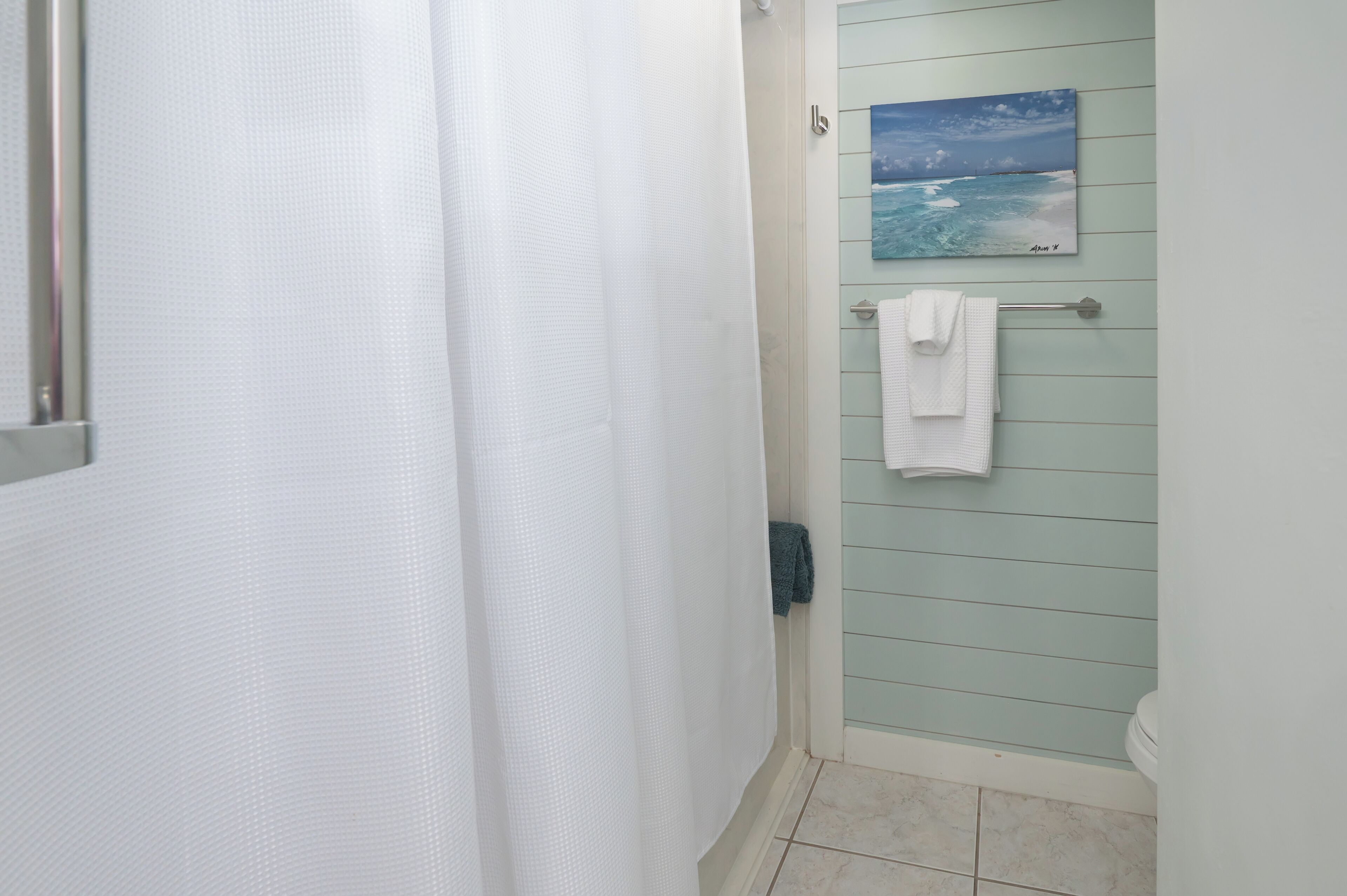 A bathroom with a shower curtain, a towel rack with two towels, and a painting of the ocean on the wall.