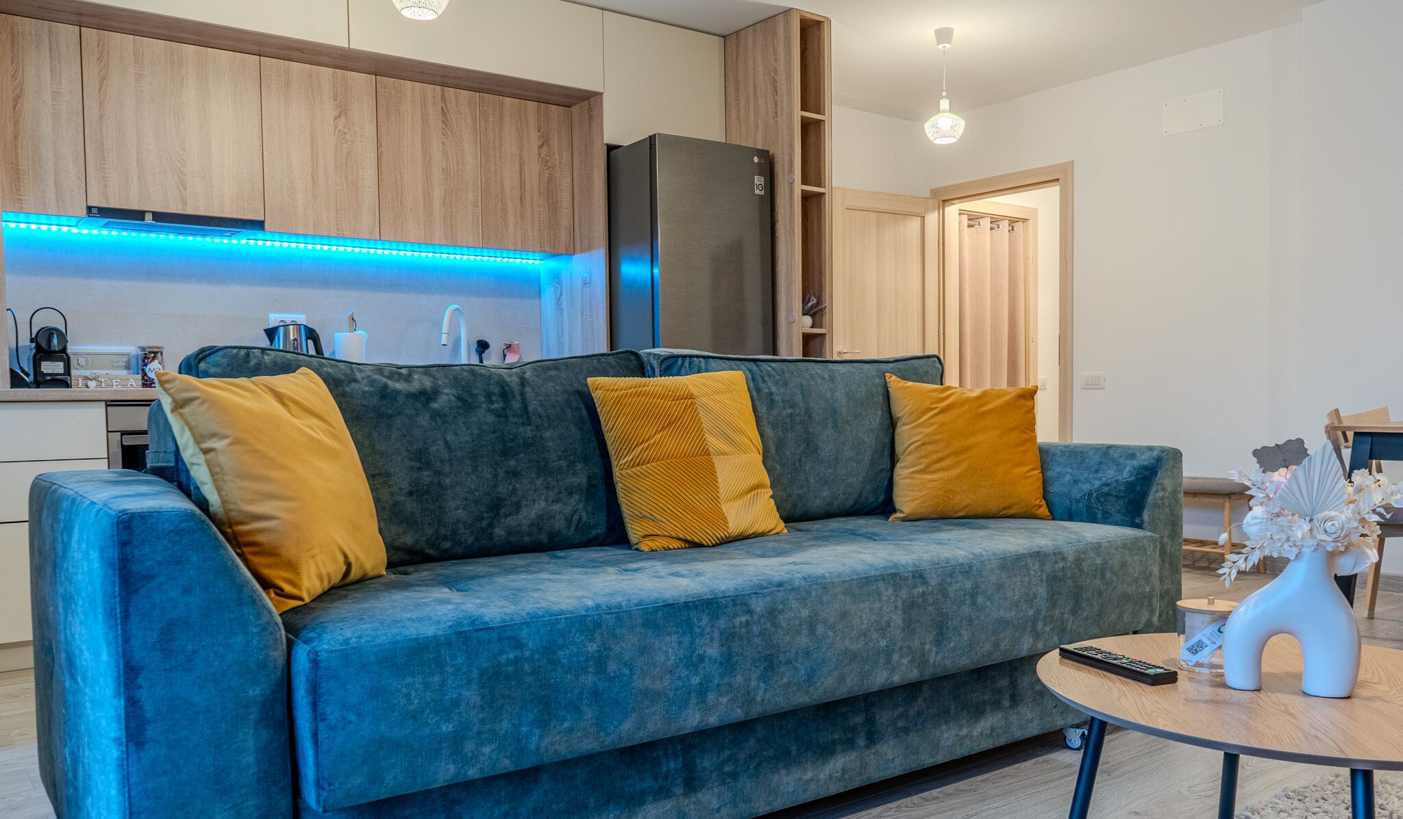 Una sala de estar moderna con un sofá azul, armarios de madera, una zona de cocina y una mesa de comedor.