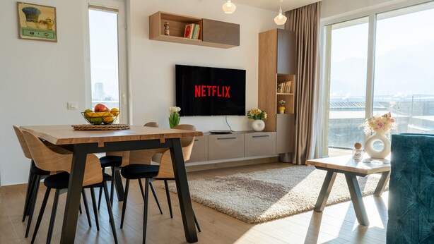 A modern living room with a dining area, a flat-screen TV displaying 'NETFLIX', a bookshelf, and a large window with a city view. - MoHost Urban Plaza (Brasov)