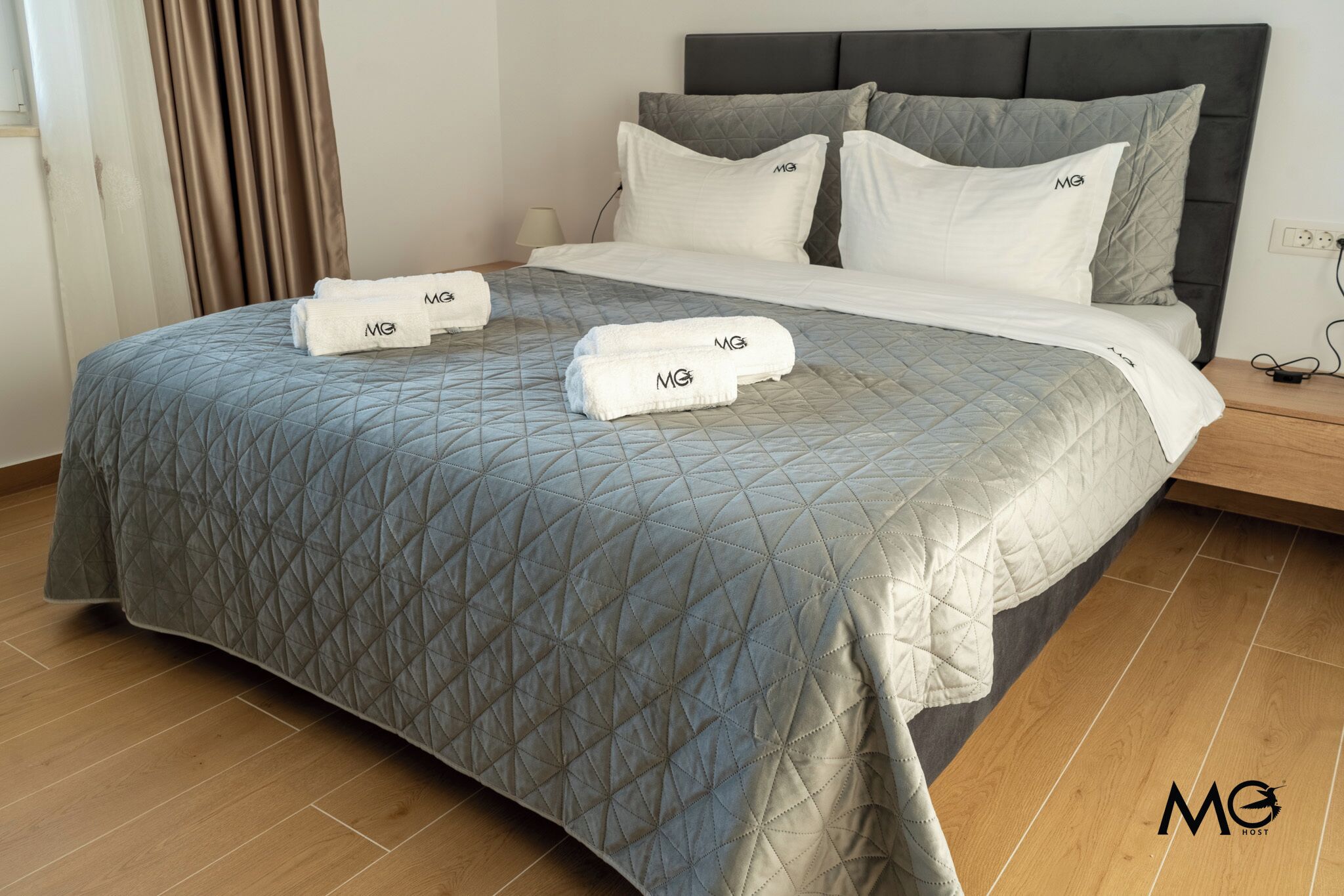 Una cama bien hecha con una colcha gris acolchada, dos almohadas con el logo de MG y dos toallas dobladas con el mismo logo.
