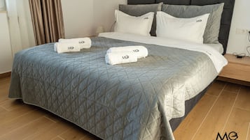 Una cama bien hecha con una colcha gris acolchada, dos almohadas con el logo de MG y dos toallas dobladas con el mismo logo.