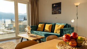 A modern living room with a teal sofa, yellow cushions, a dining table with chairs, a fruit bowl, and a cityscape view through a large window. - MoHost Urban Plaza (Brasov)