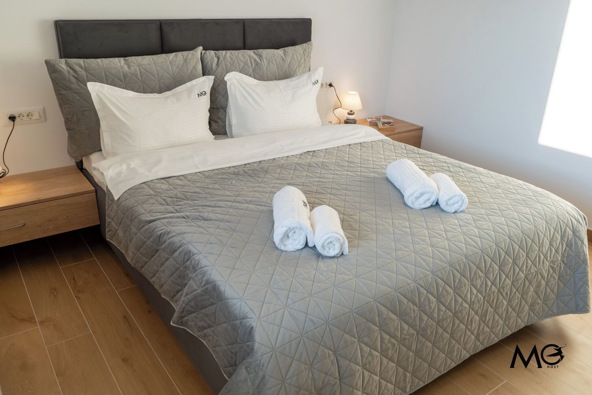 Una cama bien hecha con un edredón gris, dos almohadas y dos toallas enrolladas sobre la cama.