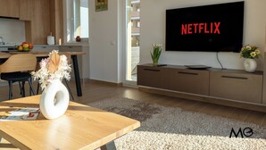 A modern living room with a flat-screen TV displaying "NETFLIX," a wooden dining table with a vase, and a kitchen in the background. - MoHost Urban Plaza (Brasov)