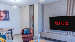 A modern living room with a large flat-screen TV displaying 'NETFLIX', a wooden dining table, a red velvet armchair, and a coffee table with a vase and remote control. - MoHost Urban Plaza (Brasov)