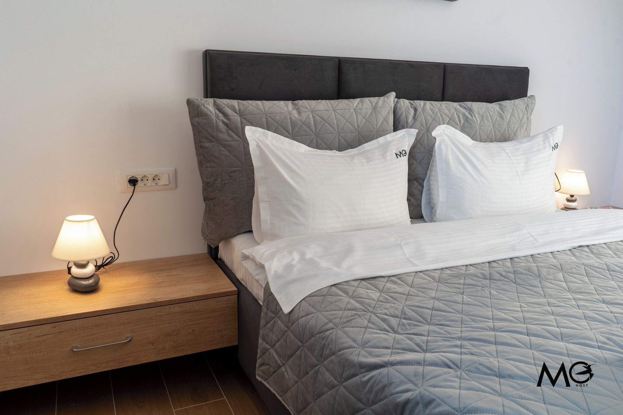 A neatly made bed with a quilted grey bedspread, two white pillows, and a grey headboard. There is a bedside table with a lamp and a power outlet on the wall.