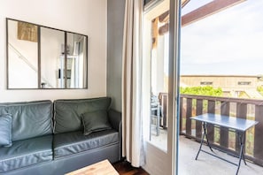 Une pièce avec un canapé en cuir noir, une petite table, un miroir et un balcon donnant sur un immeuble et de la verdure.