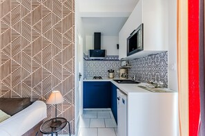 Een kleine keuken met een magnetron, kookplaat en wastafel, een eethoekje met een tafel en stoel, en een behangen wand.
