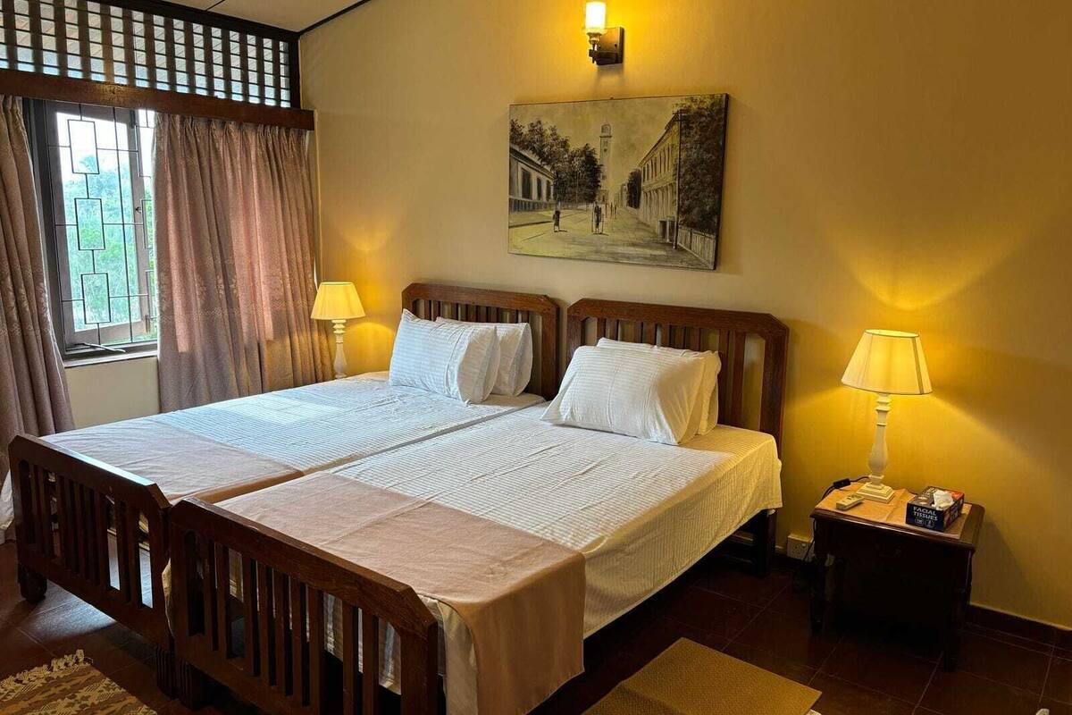 Un lit double avec une tête de lit en bois, deux tables de chevet avec des lampes, un tableau au mur et une fenêtre avec des rideaux.