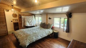 Ein Schlafzimmer mit einem Bett, Holzboden, einem Fenster, einer Wanduhr und einer Pendelleuchte.