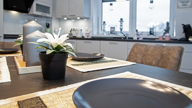 A modern kitchen with white cabinets, a dining table set with plates, a potted plant, and a window with blinds. - Above the Rooftops of Grasberg's City (Grasberg)