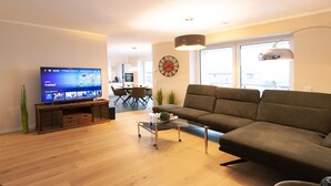 A modern living room with a flat-screen TV, a sofa, a glass coffee table, and a dining area in the background. - Above the Rooftops of Grasberg's City (Grasberg)