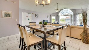 Ein Essbereich mit großem Tisch, Stühlen und einer Chandelüster. Auf dem Tisch stehen Blumentöpfe und im Hintergrund ist eine Couch zu sehen.