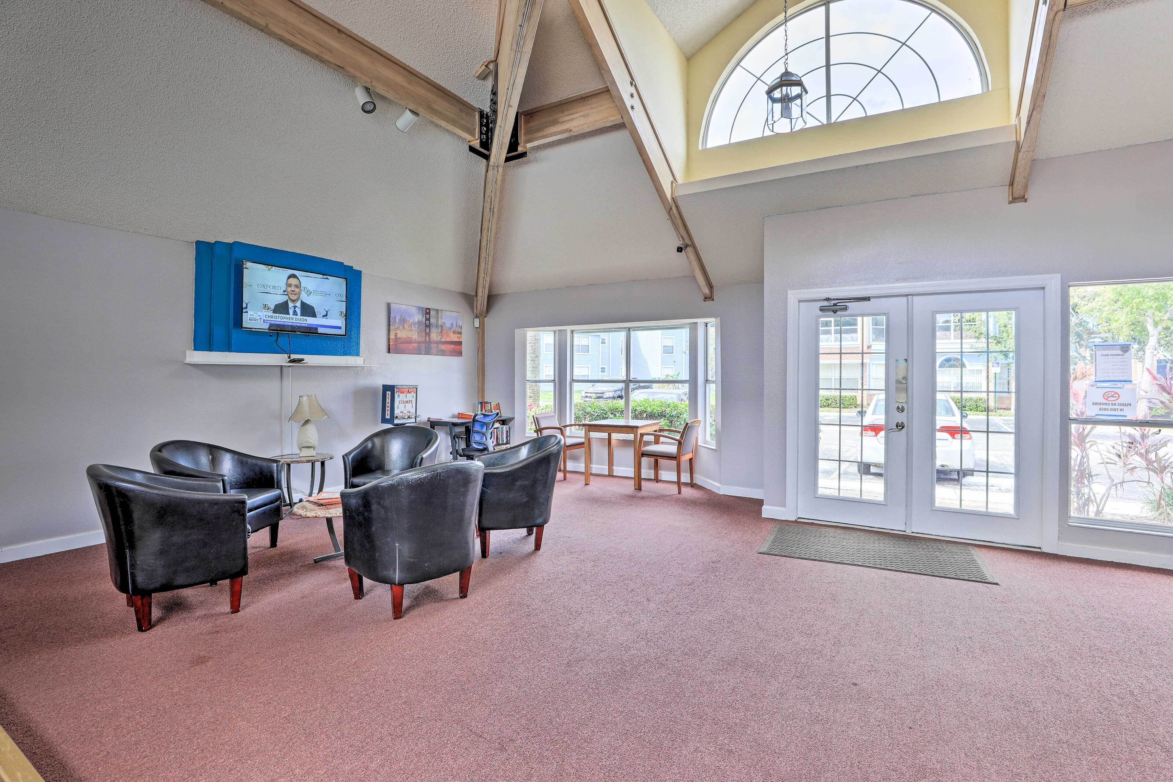 A spacious room with a high ceiling, a television mounted on the wall, a seating area with leather chairs, a small table, and a large window providing natural light.