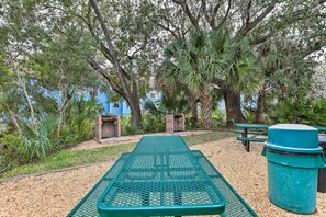 Ein Picknickplatz mit einem Metalltisch und Bänken, umgeben von Bäumen und einem blauen Gebäude im Hintergrund.