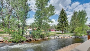 A river with a rocky bank and trees, a wooden deck, and a building in the background. - Private Hot Tub - Riverfront Cabin In Estes Park! (Estes Park)
