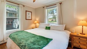 A neatly made bed with a green blanket, two bedside lamps, a wooden nightstand, and a window with curtains. - Private Hot Tub - Riverfront Cabin In Estes Park! (Estes Park)