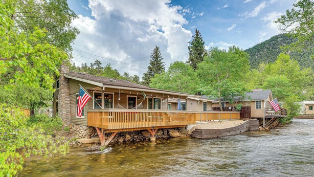 A riverside cabin with a wooden deck and American flags, surrounded by lush greenery and a mountain backdrop. - Private Hot Tub - Riverfront Cabin In Estes Park! (Estes Park)